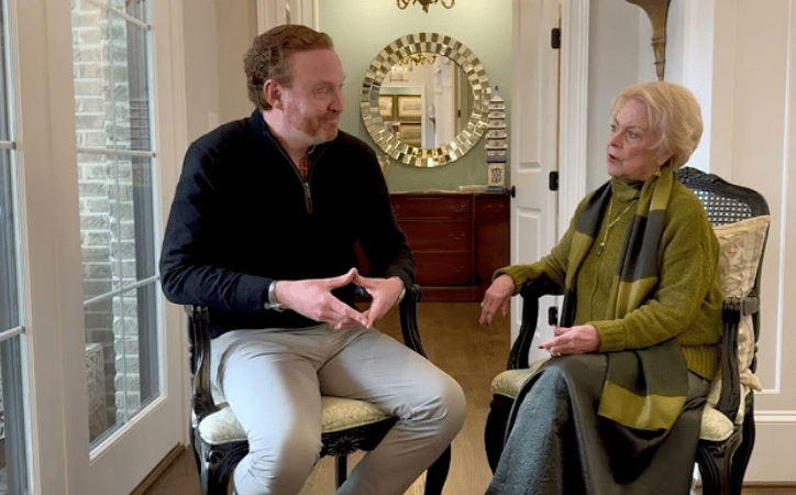 A man interviews an older woman seated in an upholstered chair indoors.