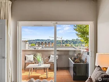 Sliding glass doors open onto a furnished balcony overlooking Seattle rooftops and green hills.