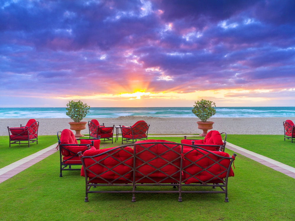 Beachfront lawn lounge with red-cushioned seating facing the ocean at sunset.