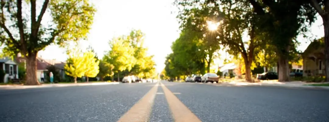 Residential street with mature trees, homes, and parked cars in warm evening sunlight.