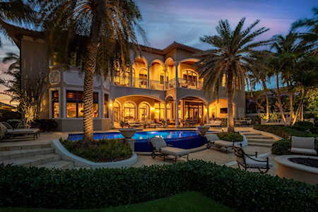 Two-story Mediterranean-style home glowing at dusk above a backyard pool framed by palms.