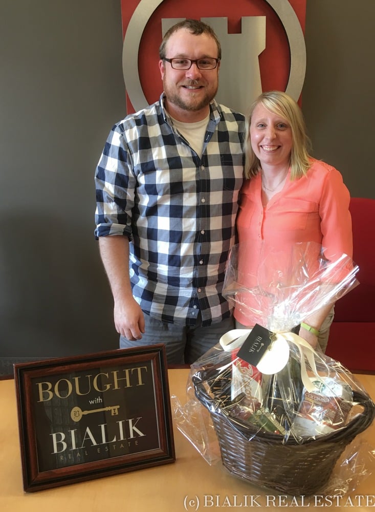 A smiling couple poses with a home-purchase sign and gift basket.