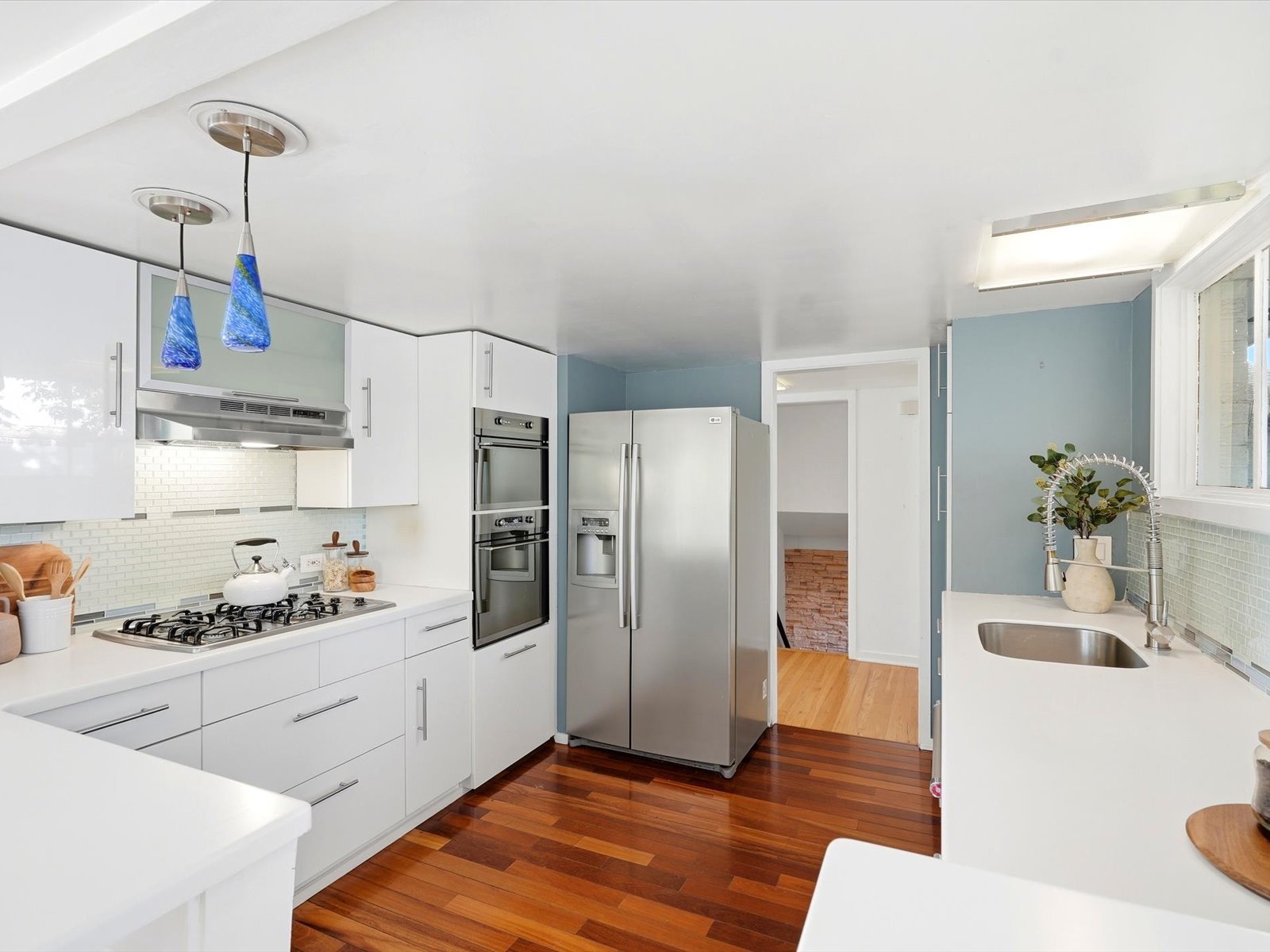 Bright white kitchen with stainless steel appliances, blue pendant lights, and warm hardwood floors.