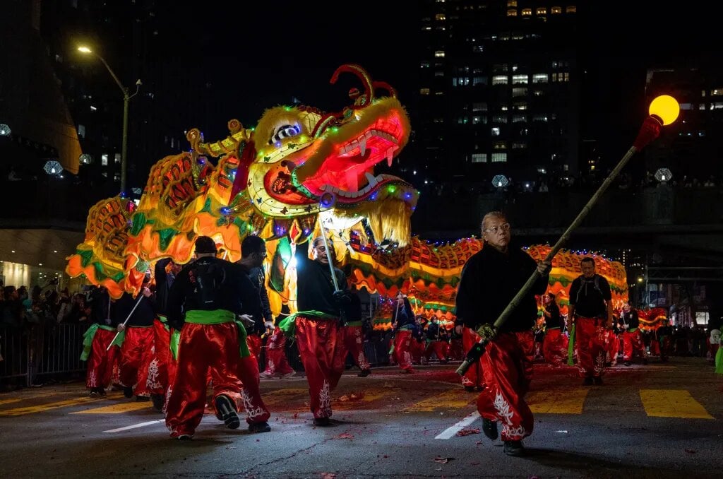 A vibrant, illuminated dragon dance performance at night during a Chinese New Year parade. Performers dressed in red and black guide the long, colorful dragon through the city streets, with glowing lights and skyscrapers in the background. A man in the foreground carries a staff with a glowing orb, leading the procession.