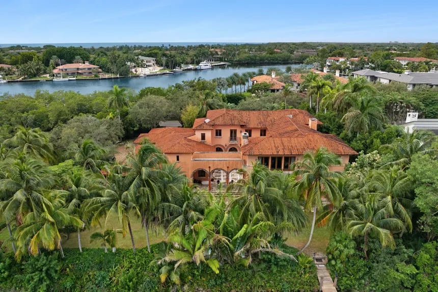 Peach-colored waterfront home with a red tile roof, palms, and a canal behind it.