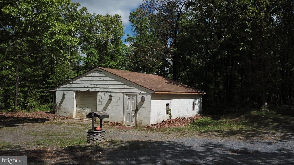 White masonry outbuilding with a brown roof beside a gravel drive in a wooded clearing.