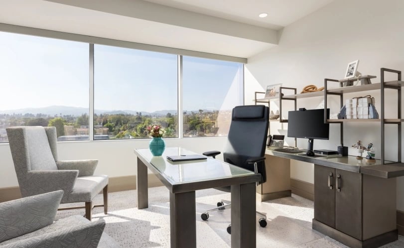 Furnished medical office with a glass-top desk and guest chairs beside large windows.