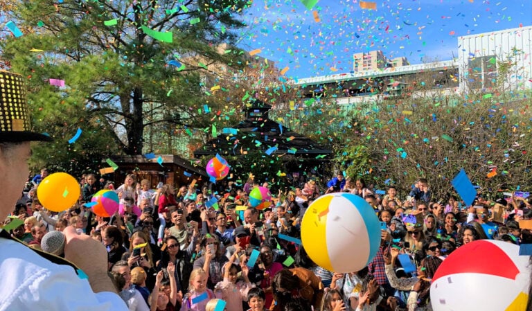 Crowd celebrates outdoors beneath falling confetti as colorful beach balls bounce overhead.