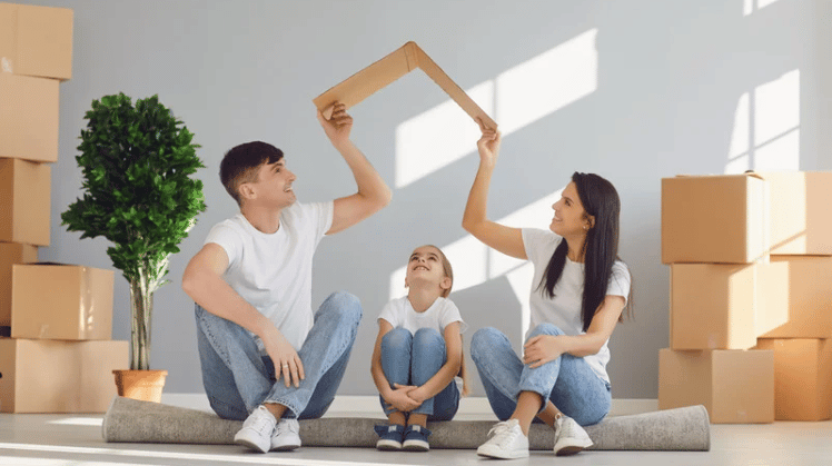 Family sits among moving boxes as the parents form a roof shape above their child.