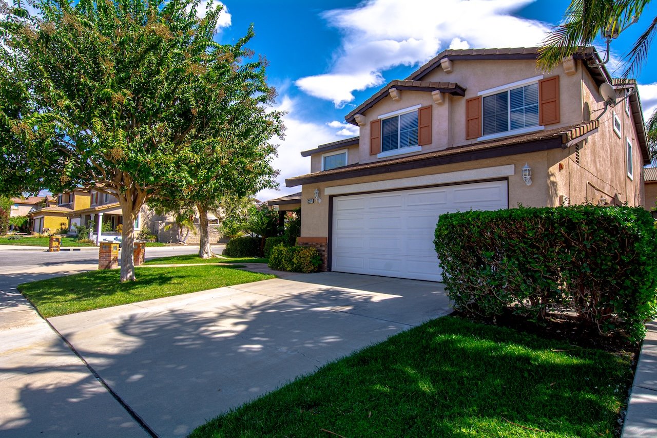 Two-story stucco home with a tiled roof, white garage door, and mature trees along the driveway.