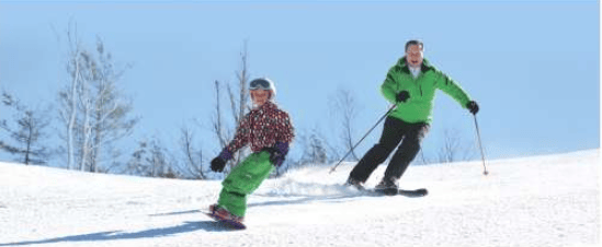 Two skiers descend a sunny snow-covered slope, one in a green jacket and the other in a patterned jacket.