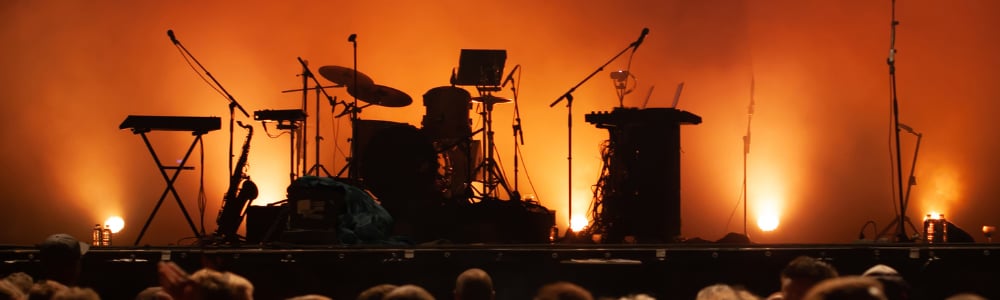 A stage setup with musical instruments, including drums and keyboards, illuminated by orange lights. Audience visible in the foreground.