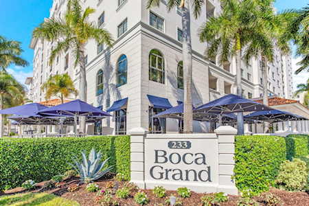 Boca Grand condominium exterior with a stone entrance sign, palm trees, and blue patio umbrellas.