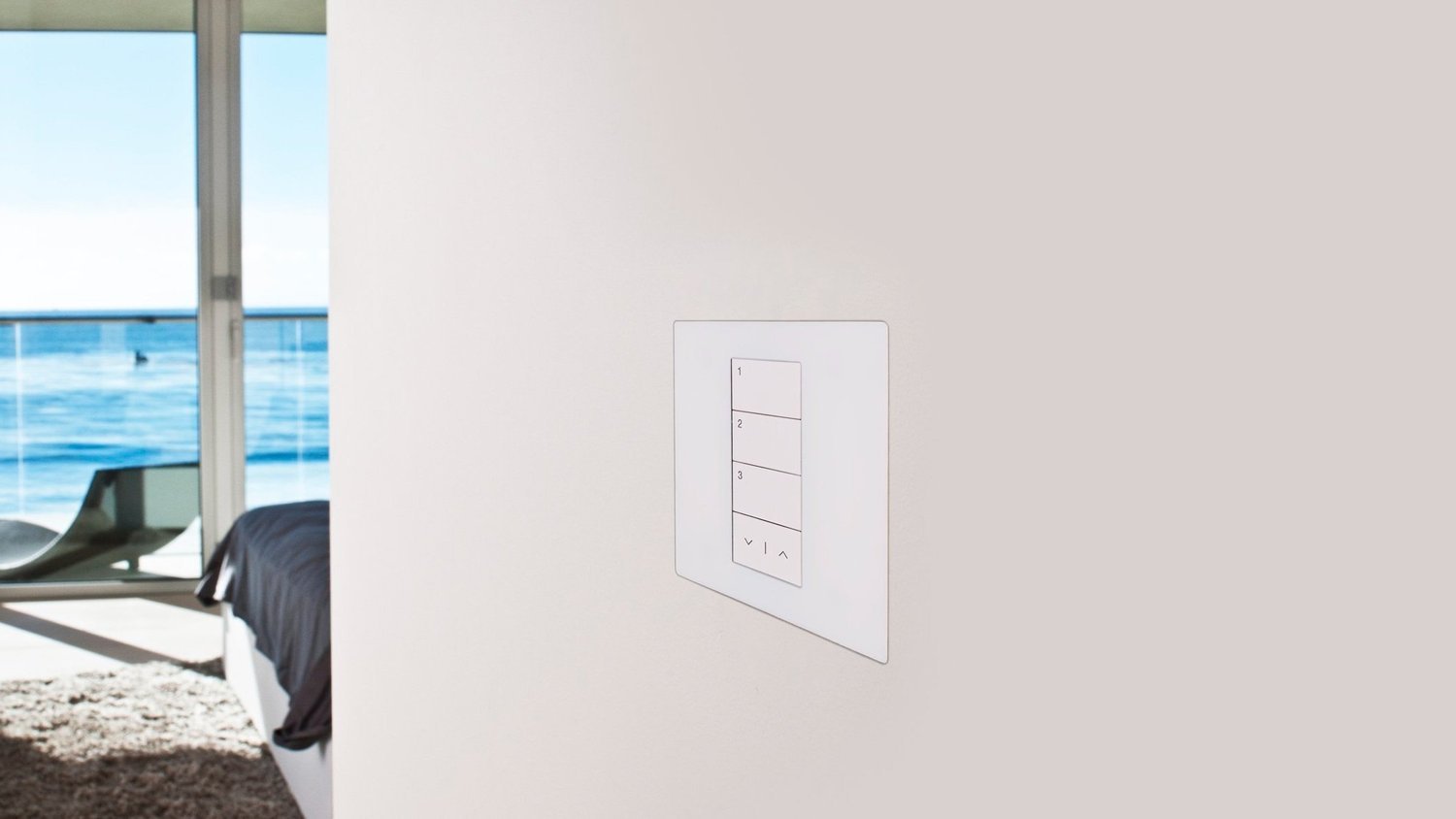 A white keypad set flush into a pale wall, with no raised plate, next to an open glass door looking out to the ocean.