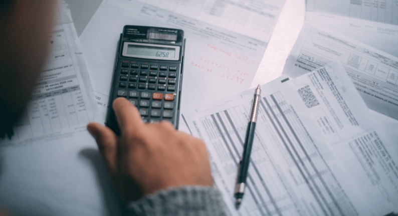 A hand uses a calculator among utility bills and statements, with a pen on the paperwork.