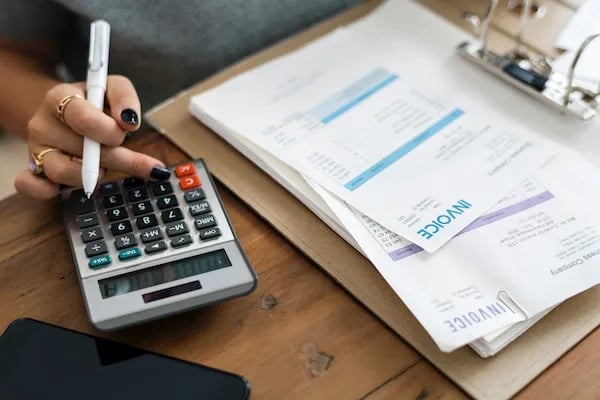 Hand calculating bills beside invoices on a wooden desk.