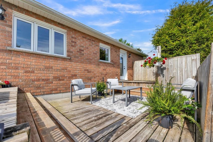 Wooden deck patio beside a brick home, furnished with lounge chairs and surrounded by potted plants.