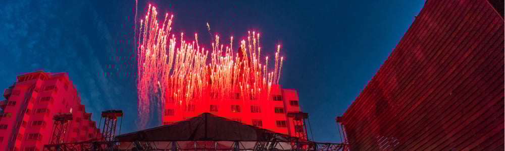 Fireworks burst in vibrant colors above buildings during an evening event, with a dramatic sky backdrop.