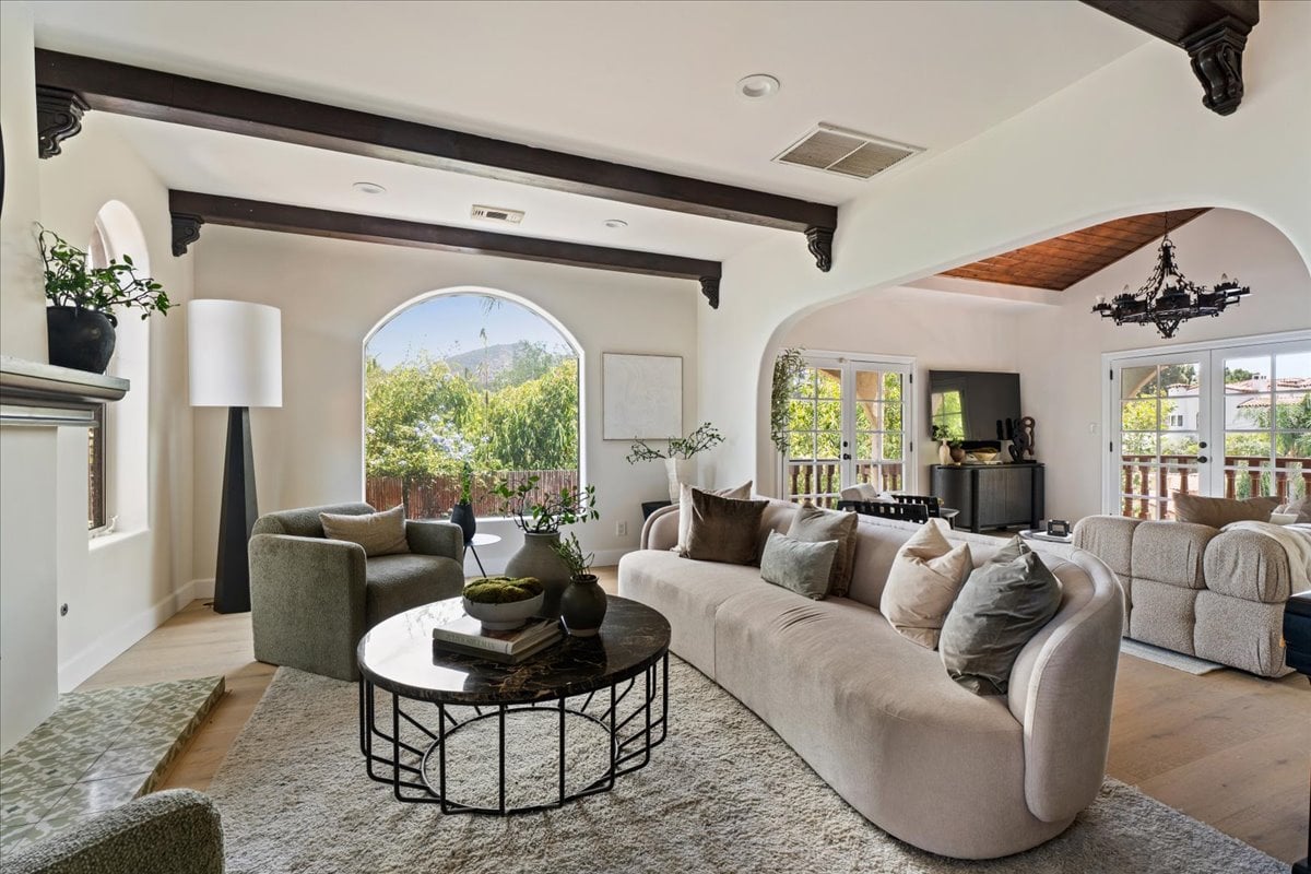 Furnished living room with cream sofas, dark ceiling beams, and arched openings to a sunny patio.