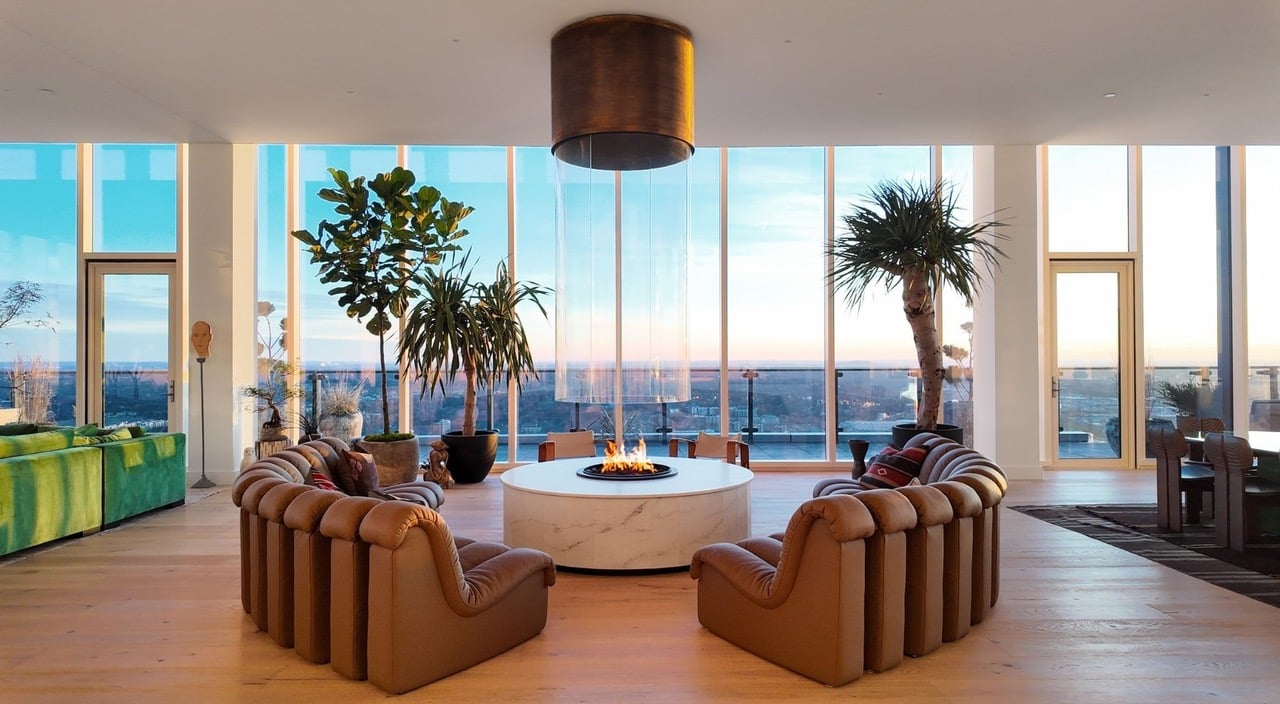 Modern living room with tan sofas around a fire table and floor-to-ceiling windows overlooking water and the city.