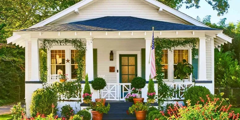 White cottage with a covered porch, American flag, and colorful flower garden.