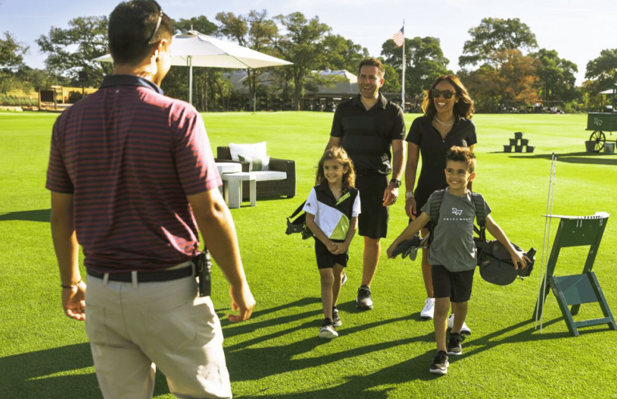 Family carrying golf bags across a sunny golf course lawn toward another person.