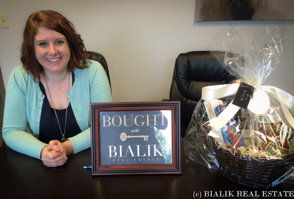Rebecca smiles beside a Bialik Real Estate closing sign and gift basket.