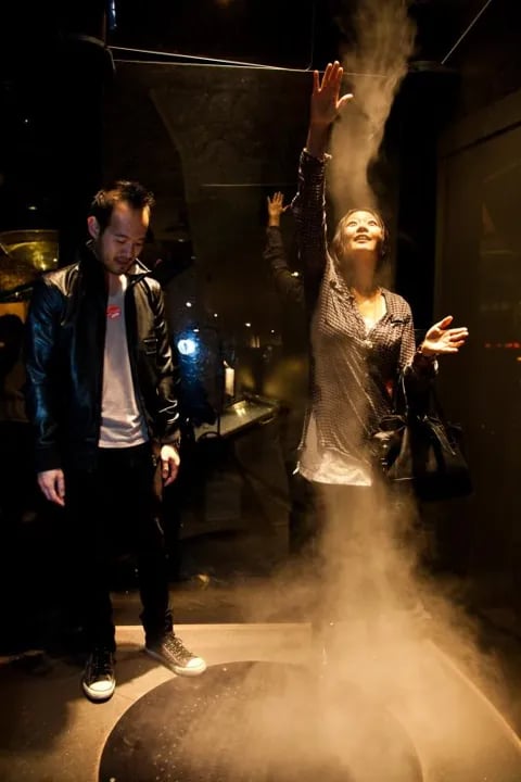 Color photo of a couple at an After Dark Thursday Nights event, standing in a dimly lit science exhibit space; the woman raises her arms in delight as a dramatic plume of swirling fog rises from a floor vent around her, while the man smiles beside her in a black leather jacket.