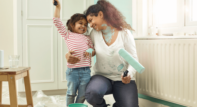 A mother and daughter happily paint a room, both smiling widely. The child holds a brush, and the mother kneels with a paint roller. Walls have splashes of light blue.