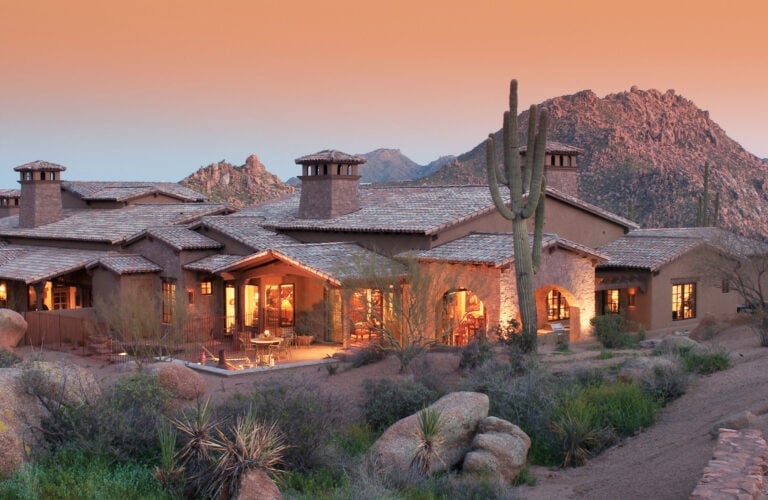 Southwestern-style residence glowing at dusk, with a saguaro cactus and rocky mountains behind it.