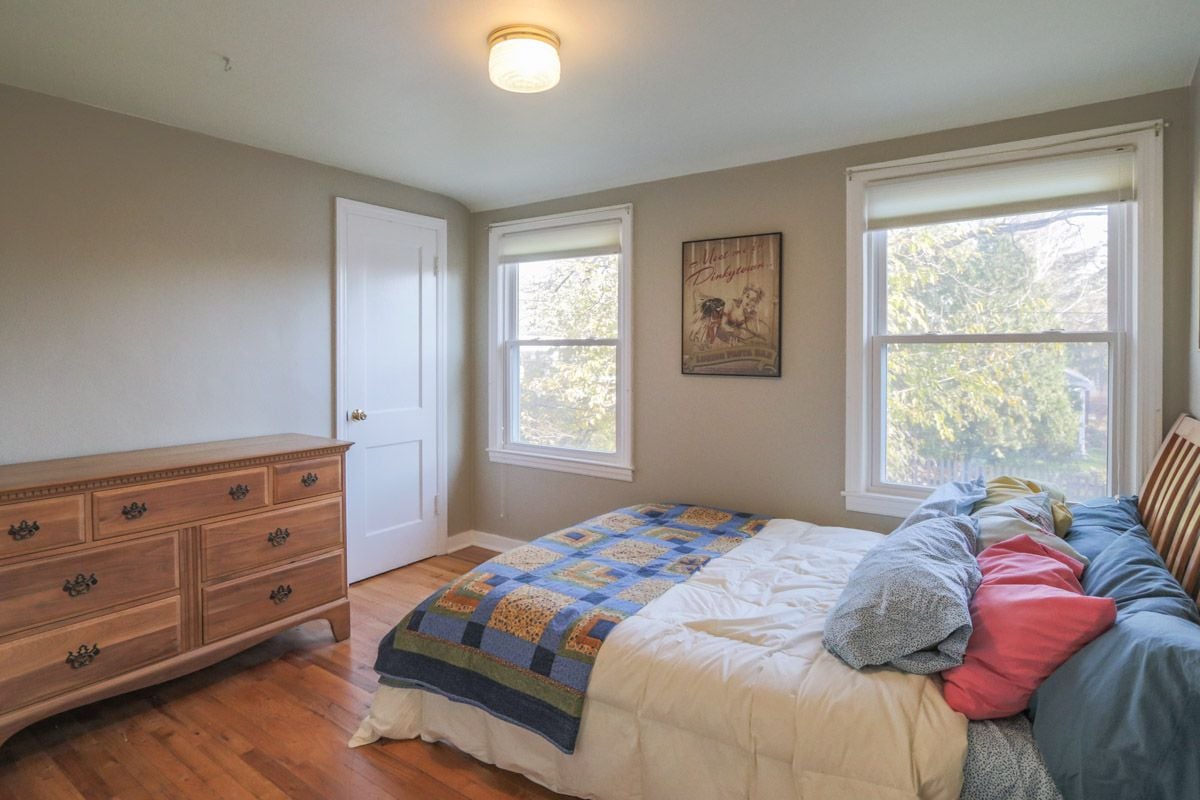 Bedroom with a blue patchwork quilt, wooden dresser, and two sunlit windows.