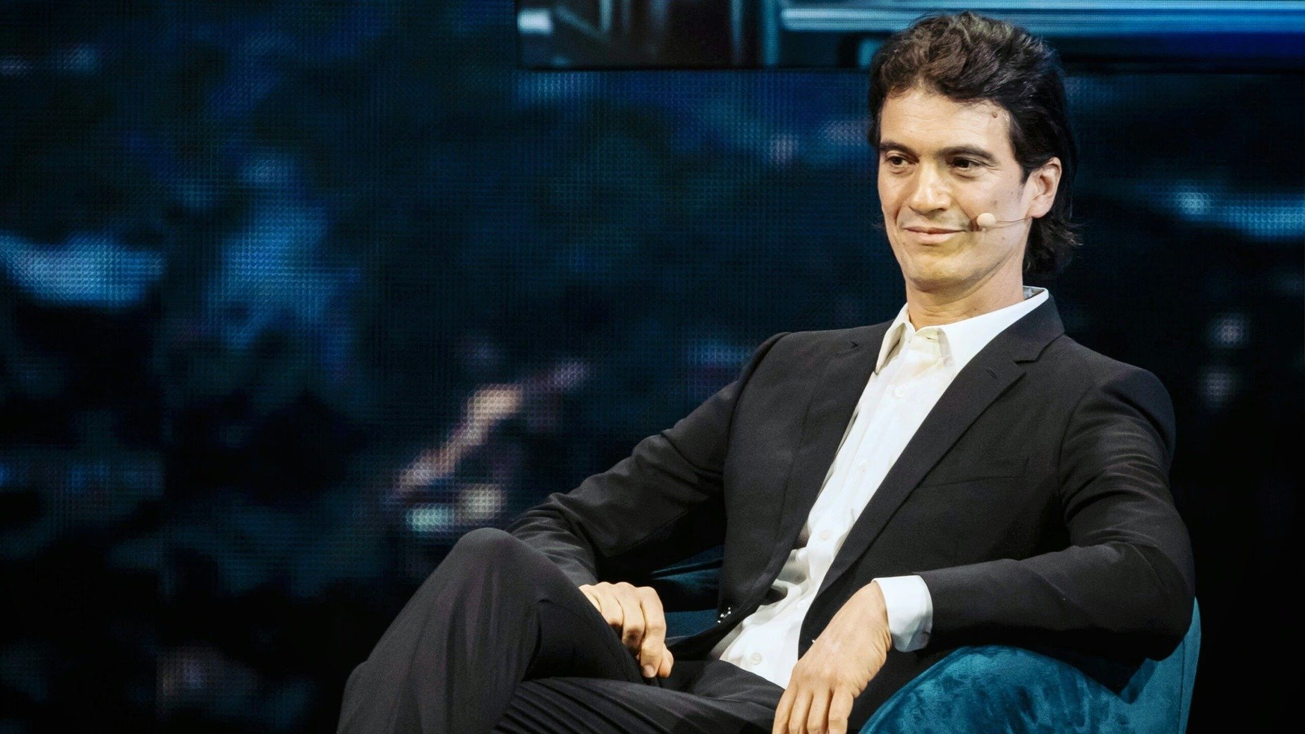 Dark-haired man in a black suit seated in a teal chair onstage.