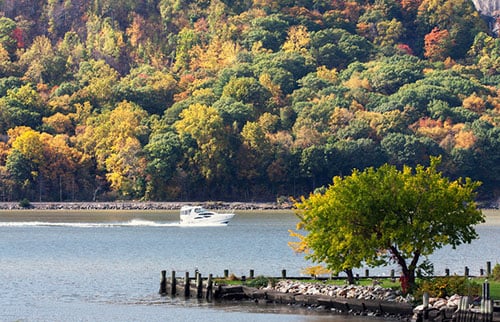 Autumn riverbank with colorful wooded hills, a small boat, and a tree on a rocky point.