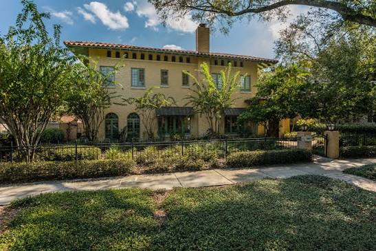 Two-story yellow stucco home with a tile roof, arched windows, and a landscaped yard behind a metal fence.