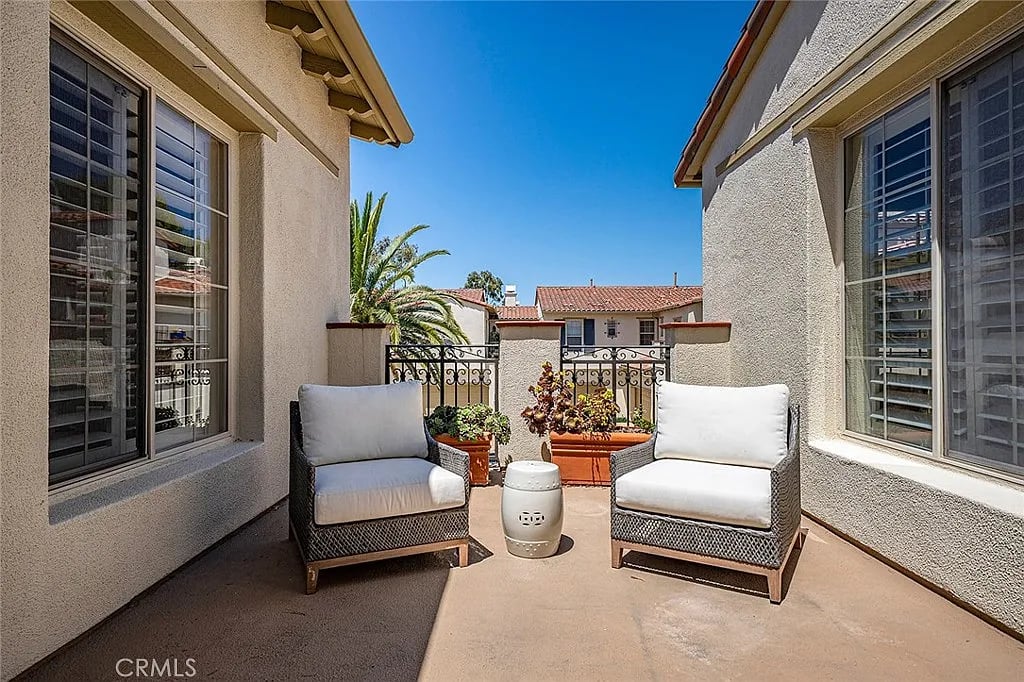 Patio with two cushioned wicker chairs, a white side table, potted plants, and a decorative railing.
