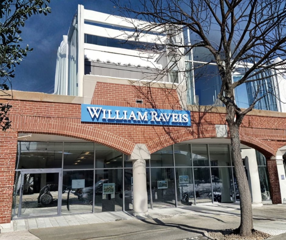 William Raveis real estate office with a brick facade, glass storefront, and bare trees.