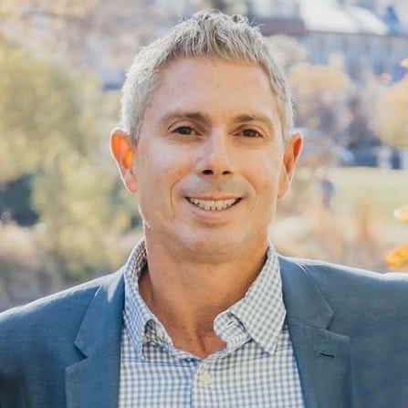 Smiling man in a blue blazer and checked shirt against a blurred outdoor background.