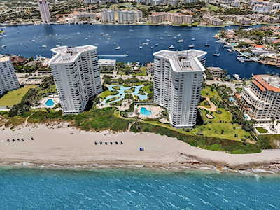 Aerial view of beachfront condominium towers between turquoise ocean and a boat-filled waterway.