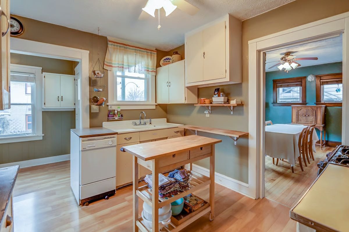 Kitchen with a rolling wooden worktable, white cabinets, and a view into a teal dining room.