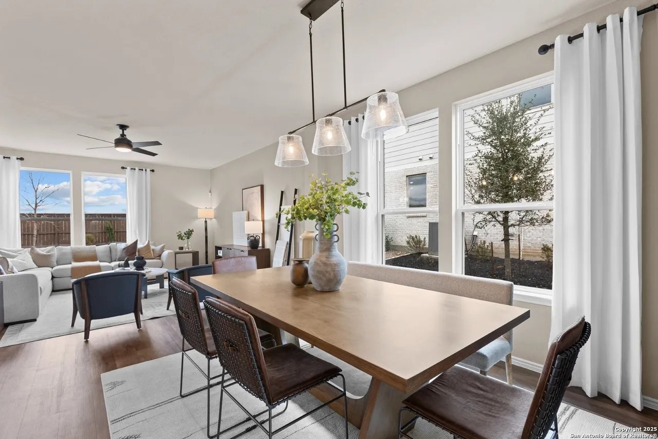 Open-plan dining and living area with a wood table, woven chairs, and tall curtained windows.