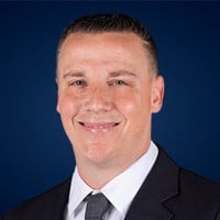 Smiling man in a dark suit and tie against a dark blue background.