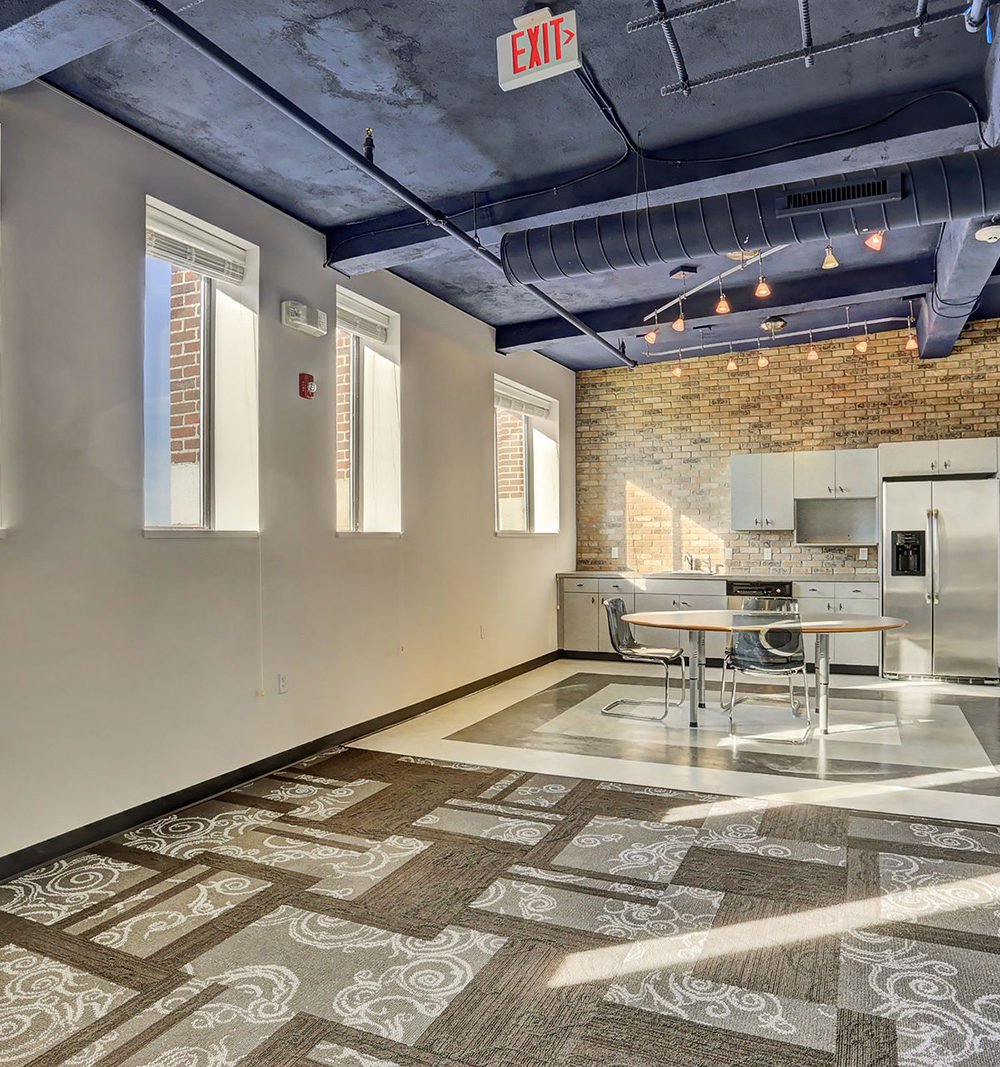 Common room with a kitchenette, round dining table, tall windows, and exposed ceiling ductwork.