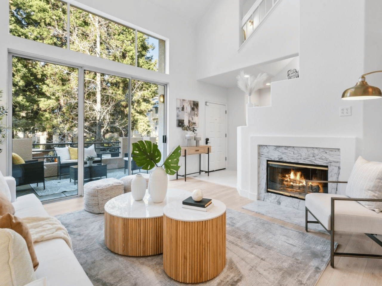 A bright, modern living room featuring a fireplace, large windows, and stylish furniture, with a view of greenery outside.