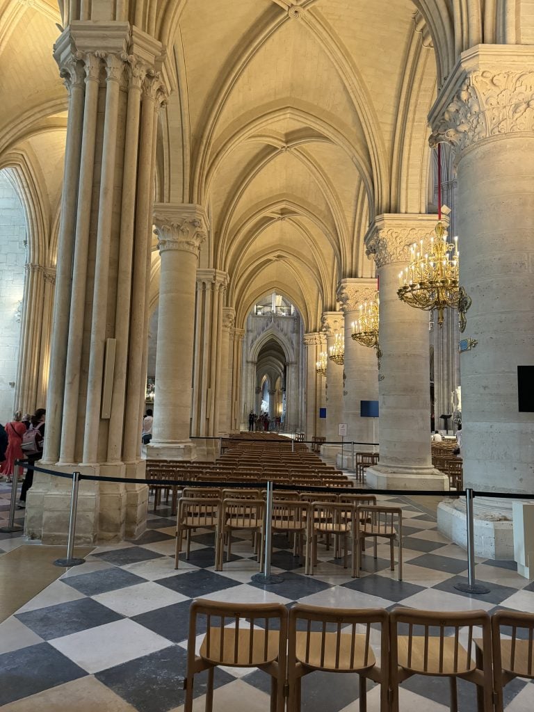 Gothic cathedral interior with soaring stone vaults, chandeliers, and rows of wooden chairs.