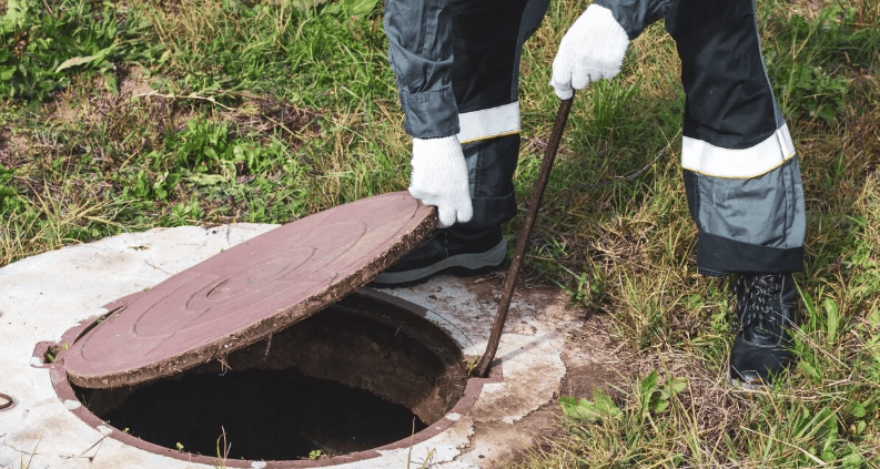 Worker lifts a septic tank cover in a grassy area.