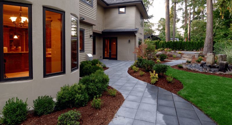 Gray-sided home with a curved paved walkway, landscaped garden beds, and warm-lit windows.