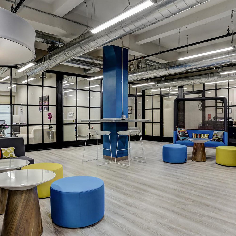 Communal lounge with colorful seating, glass-partitioned rooms, and exposed ceiling ducts.