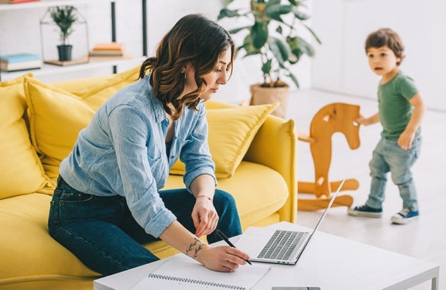 A woman works on her laptop beside a yellow sofa as a young child stands nearby.