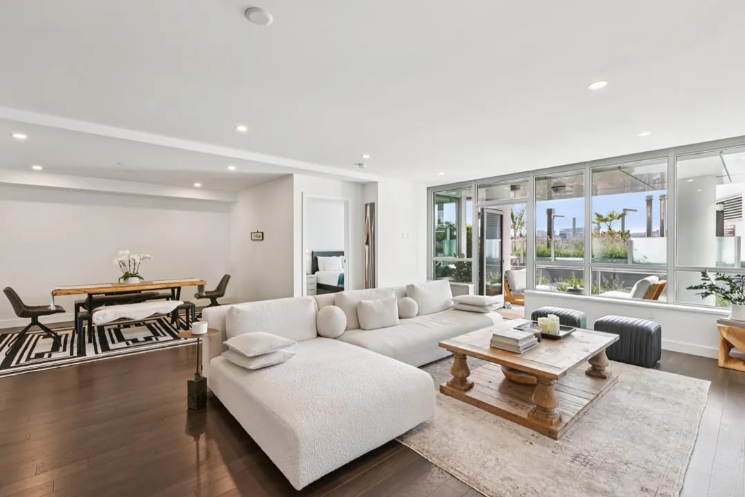 A bright, modern open-plan living and dining room with dark wood floors. The living area has a large white sectional sofa and a rustic wood coffee table on a light rug, while the dining area features a natural wood table with a bench and black chairs on a geometric rug. Floor-to-ceiling windows look out onto a balcony.