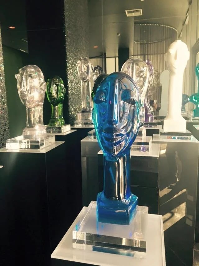 Blue glass face sculpture displayed among colorful glass figures on clear pedestals.